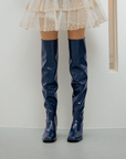 Front view of a model wearing Lisa Iris Blue boots by ACQUO of Sweden. The image highlights iris blue knee-high rubber boots with a sleek reflective finish and square toe silhouette, styled with a layered ivory tulle skirt in a minimalist indoor setting.