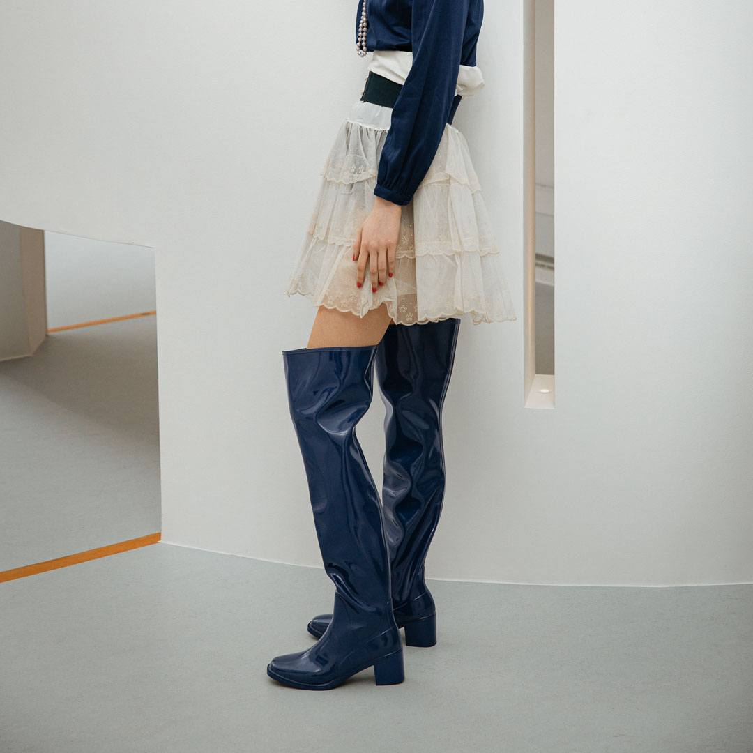 Side view of a model wearing Lisa Iris Blue boots by ACQUO of Sweden. The image features iris blue knee-high rubber boots with a sleek reflective finish and block heel, styled with a layered ivory tulle skirt, navy blouse, and pearl necklace in a minimalist indoor setting.
