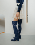 Side view of a model wearing Lisa Iris Blue boots by ACQUO of Sweden. The image features iris blue knee-high rubber boots with a sleek reflective finish and block heel, styled with a layered ivory tulle skirt, navy blouse, and pearl necklace in a minimalist indoor setting.