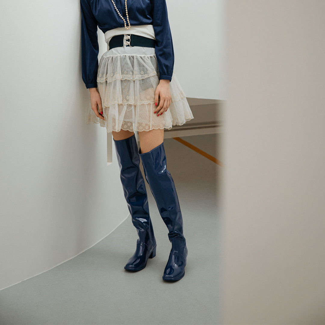Model standing between curved white walls wearing Lisa Iris Blue boots by ACQUO of Sweden. The image features iris blue over-the-knee rubber boots with a sleek reflective finish and block heel, styled with a navy blouse, layered ivory tulle skirt, and pearl necklace in a minimalist indoor setting.