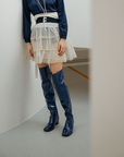 Model standing between curved white walls wearing Lisa Iris Blue boots by ACQUO of Sweden. The image features iris blue over-the-knee rubber boots with a sleek reflective finish and block heel, styled with a navy blouse, layered ivory tulle skirt, and pearl necklace in a minimalist indoor setting.