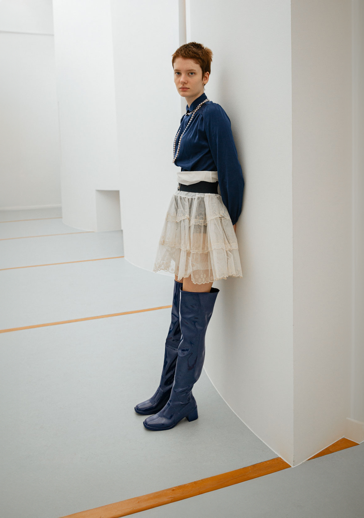 Model leaning against a curved white wall while wearing Lisa Iris Blue boots by ACQUO of Sweden. The image features iris blue knee-high rubber boots with a sleek reflective finish and block heel, styled with a navy blouse, layered ivory tulle skirt, and pearl necklace in a minimalist architectural setting.