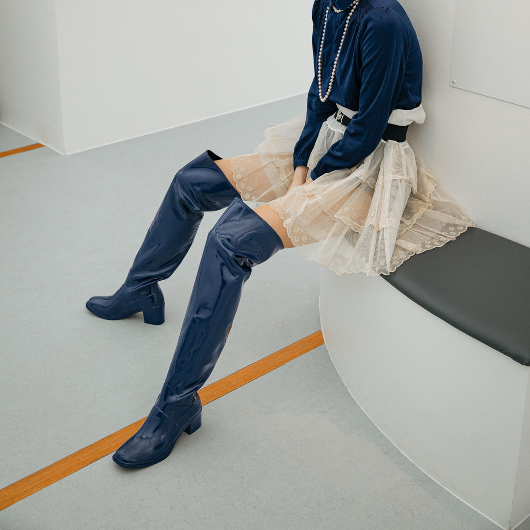 Model seated on a curved bench wearing Lisa Iris Blue boots by ACQUO of Sweden. The image features iris blue over-the-knee rubber boots with a reflective finish and block heel, styled with a navy blouse, layered tulle skirt, and pearl necklace in a minimalist indoor setting.