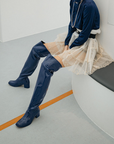 Model seated on a curved bench wearing Lisa Iris Blue boots by ACQUO of Sweden. The image features iris blue over-the-knee rubber boots with a reflective finish and block heel, styled with a navy blouse, layered tulle skirt, and pearl necklace in a minimalist indoor setting.