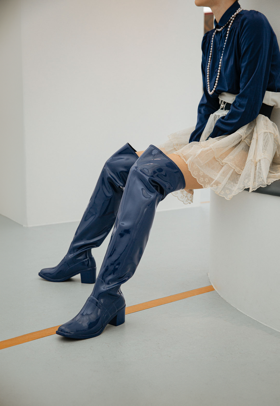 Close-up side view of a model seated on a curved bench wearing Lisa Iris Blue boots by ACQUO of Sweden. The image highlights iris blue over-the-knee rubber boots with a sleek reflective finish and block heel, styled with a navy blouse, pearl necklace, and layered ivory tulle skirt in a minimalist indoor setting