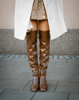 Close-up of a woman wearing Lisa Walnut Brown knee-high boots by ACQUO of Sweden with a beige patterned dress and white coat. Elegant waterproof boots in rich brown with a sleek Scandinavian silhouette styled for modern city fashion