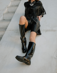Model wearing ACQUO of Sweden’s sleek shine black boots, paired with a black leather outfit with fringes, lying on a modern concrete staircase