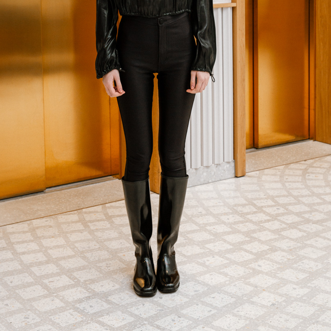 Front view of ACQUO of Sweden's sleek knee-high black rubber boots styled with fitted black trousers and a satin blouse—timeless Scandinavian craftsmanship meets contemporary fashion