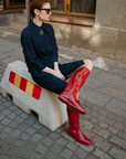 Woman sitting on a concrete barrier in a city street wearing Svea Cherry Red knee-high boots by ACQUO of Sweden with a navy textured dress and sunglasses. Waterproof boots with elegant Swedish design and contemporary Scandinavian fashion styling.