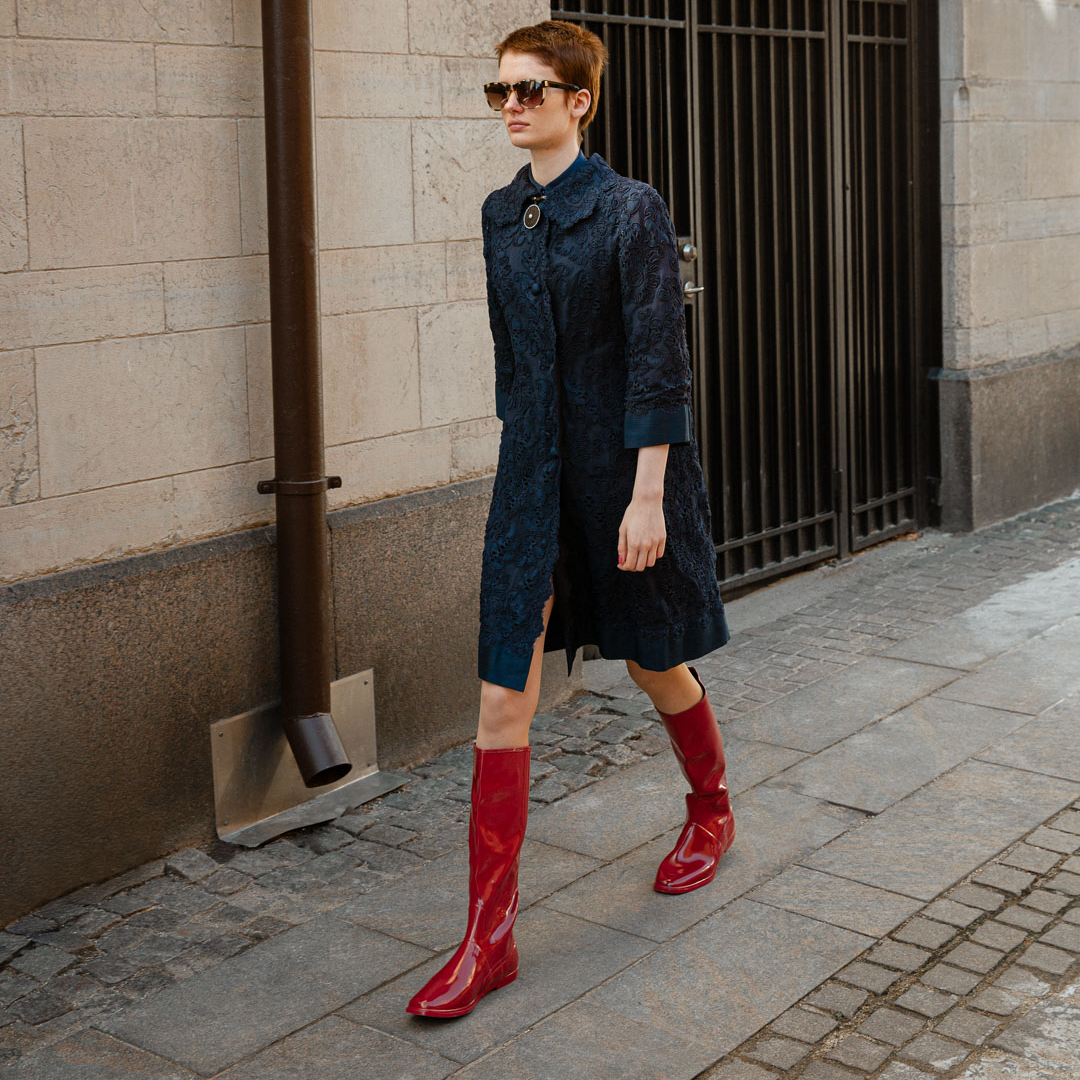 Woman walking through a city street wearing Svea Cherry Red knee-high boots by ACQUO of Sweden with a navy embroidered dress and sunglasses. Waterproof boots featuring elegant Swedish design and modern Scandinavian fashion