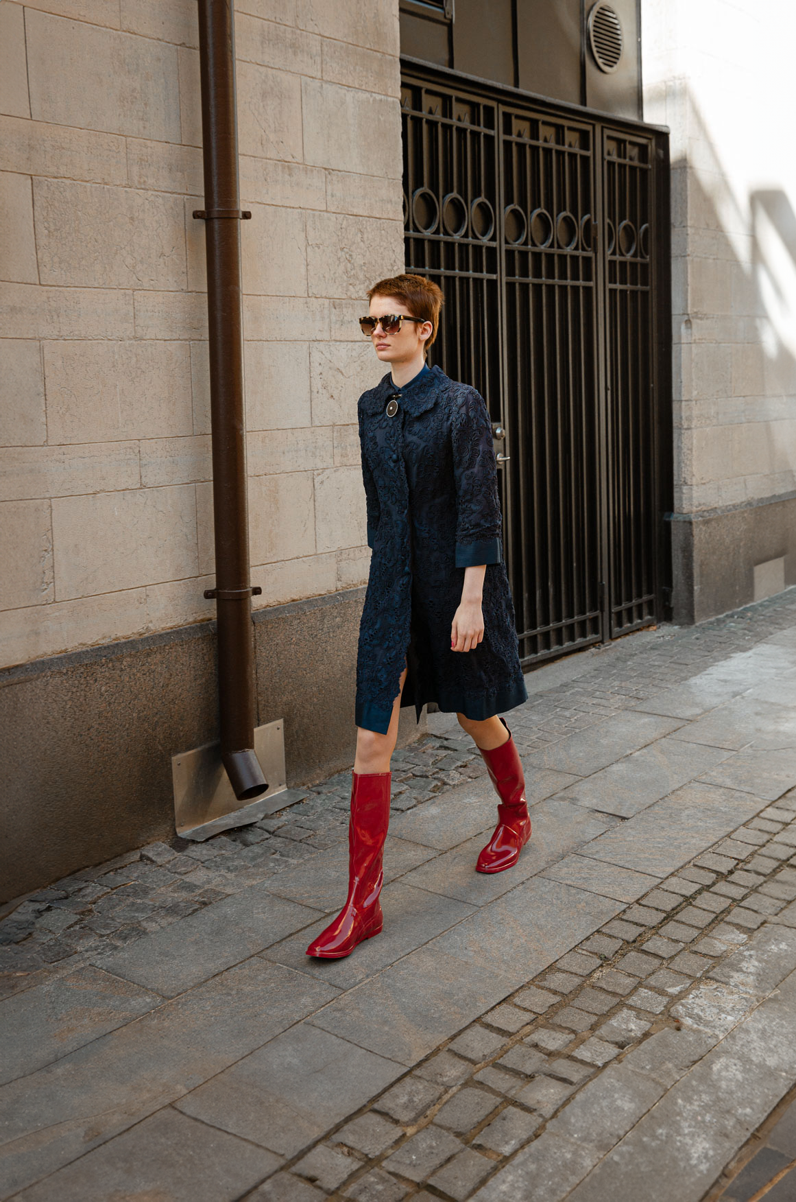 Woman walking through a city street wearing Svea Cherry Red knee-high boots by ACQUO of Sweden with a navy embroidered dress and sunglasses. Waterproof boots featuring elegant Swedish design and modern Scandinavian fashion