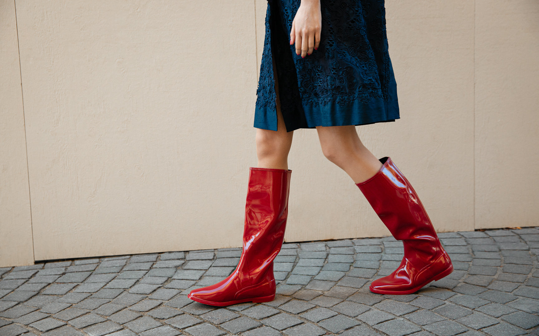 Side view of ACQUO of Sweden Svea Cherry Red knee-high natural rubber boots, styled with a navy dress in a modern Scandinavian street setting