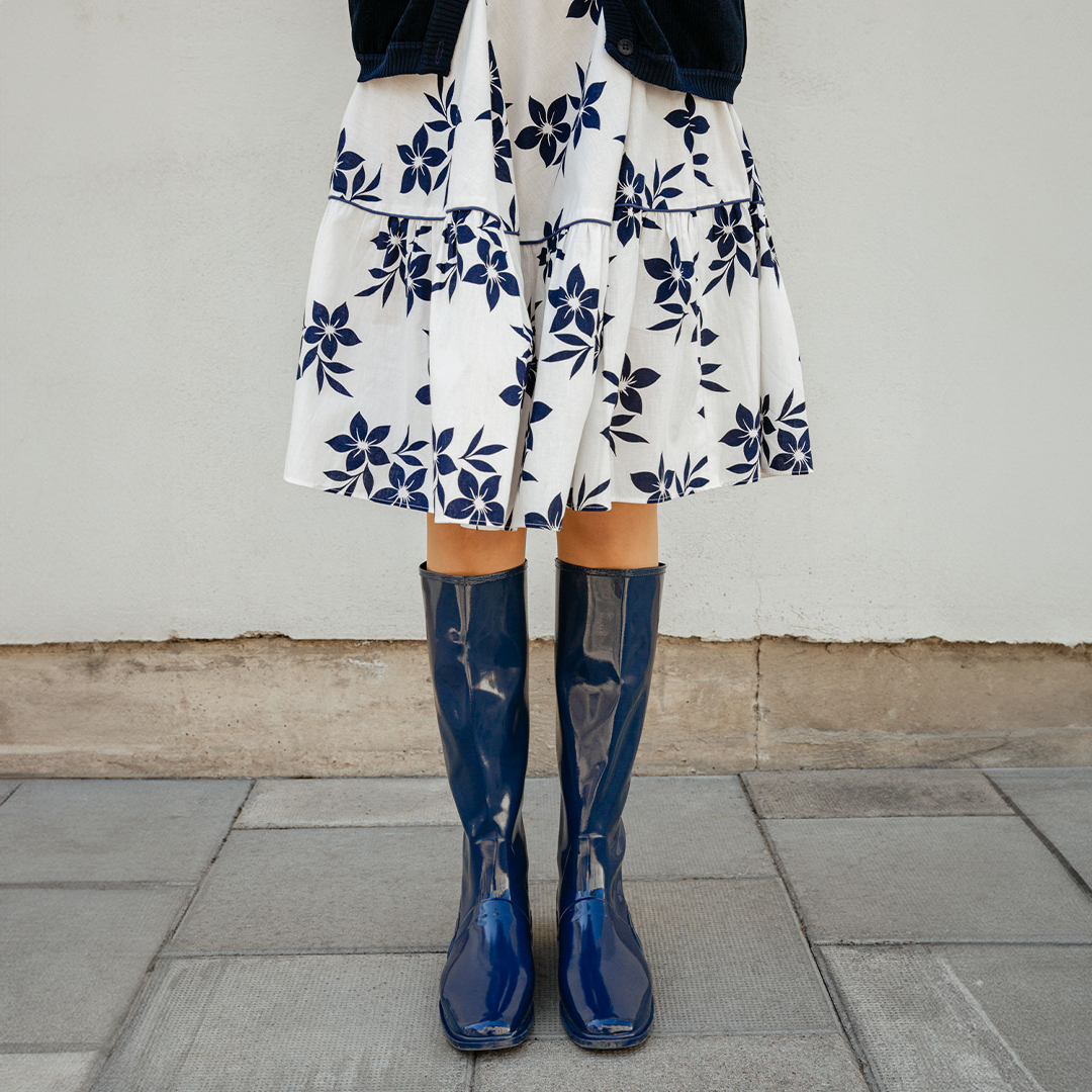 Front view of Svea Iris Blue boots by ACQUO of Sweden styled with a white floral skirt and navy cardigan. The image features iris blue knee-high rubber boots with a sleek reflective finish photographed on a city pavement against a light stone wall.