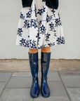 Front view of Svea Iris Blue boots by ACQUO of Sweden styled with a white floral skirt and navy cardigan. The image features iris blue knee-high rubber boots with a sleek reflective finish photographed on a city pavement against a light stone wall.