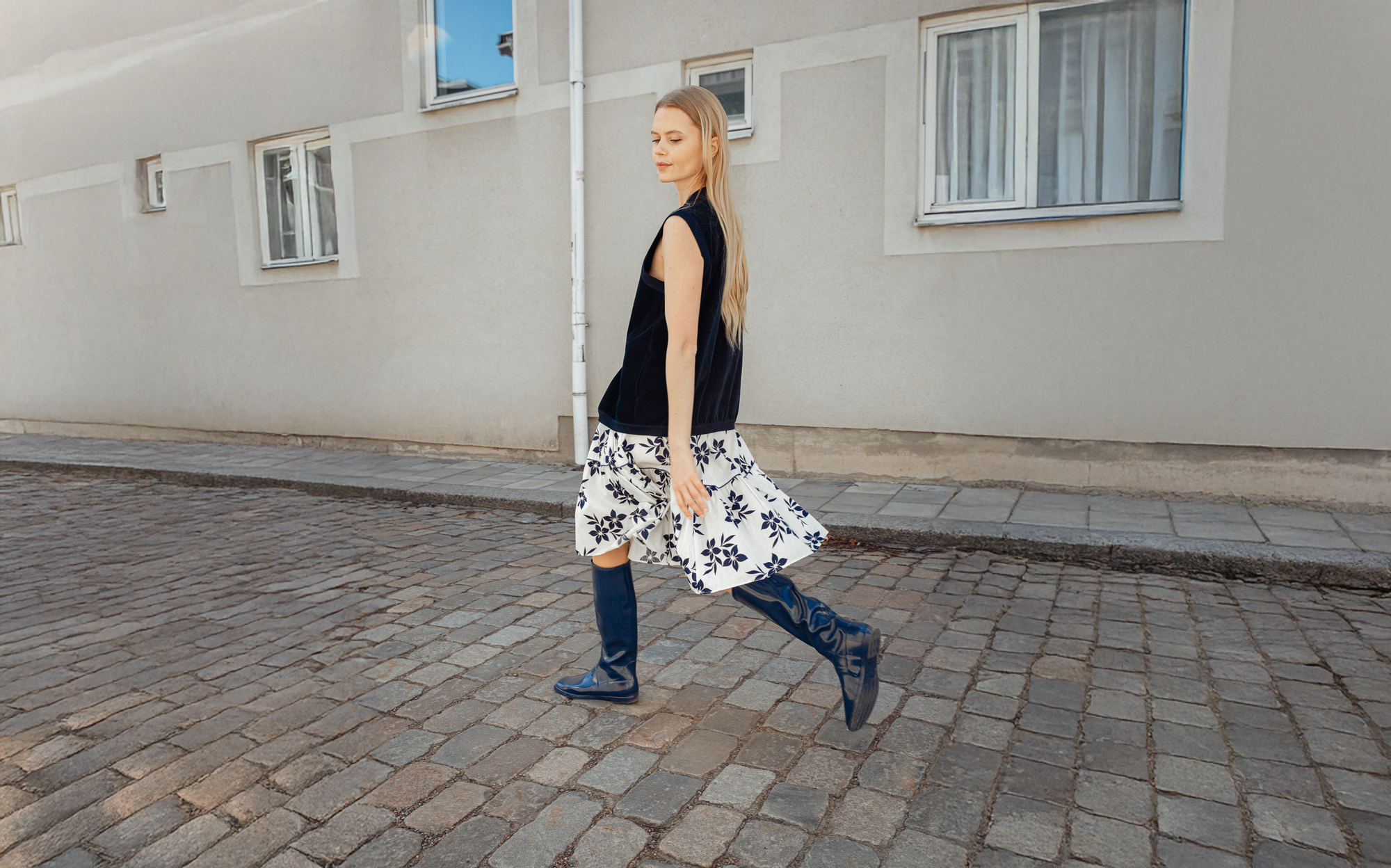 Woman walking on a cobblestone street wearing navy blue knee-high waterproof boots by ACQUO of Sweden with a floral skirt and minimalist Scandinavian styling.