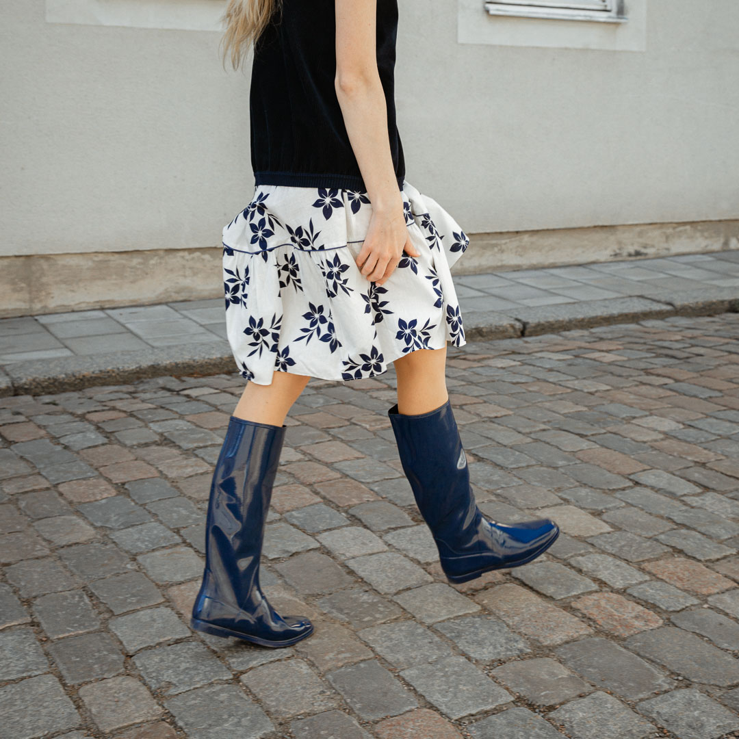 Side view of a model walking on a cobblestone street wearing Svea Iris Blue boots by ACQUO of Sweden. The image features iris blue knee-high rubber boots with a sleek reflective finish, styled with a black top and white floral skirt in an urban outdoor setting