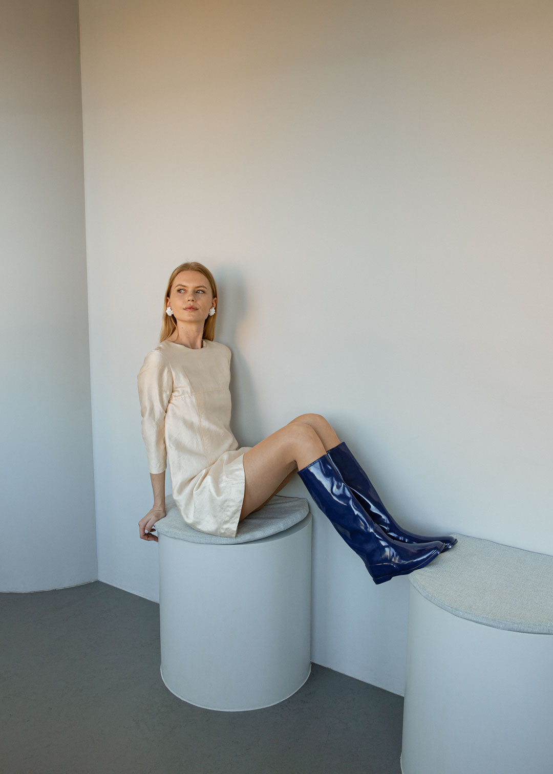 Model wearing Svea Iris Blue knee-high rubber boots by ACQUO of Sweden with a cream mini dress in a minimalist indoor setting. The elegant blue waterproof boots feature a sleek reflective finish and modern Scandinavian style.