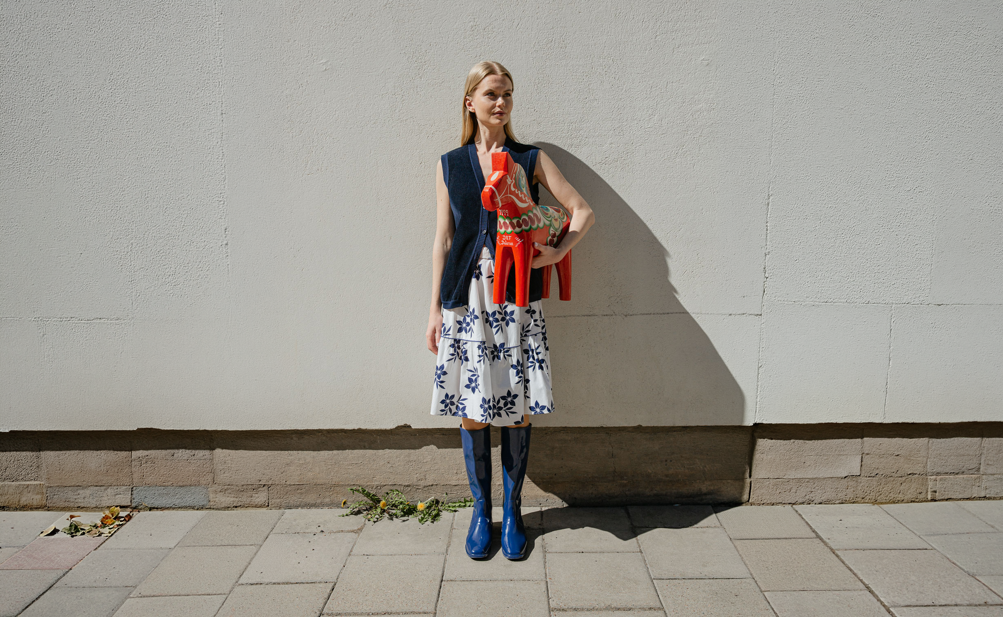 Model wearing ACQUO of Sweden Iris Blue natural rubber boots, styled with a white floral skirt and navy knit vest while holding a traditional Swedish Dala horse.