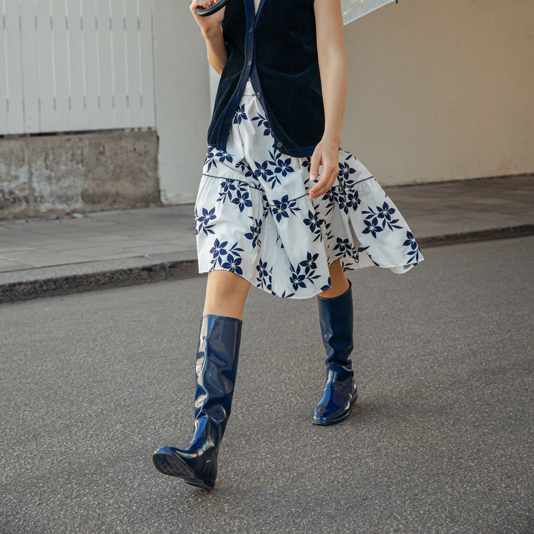 Model walking outdoors in Svea Iris Blue knee-high rubber boots by ACQUO of Sweden, styled with a white floral skirt and navy cardigan. The image highlights elegant blue waterproof boots with a sleek reflective finish in a contemporary street-style setting
