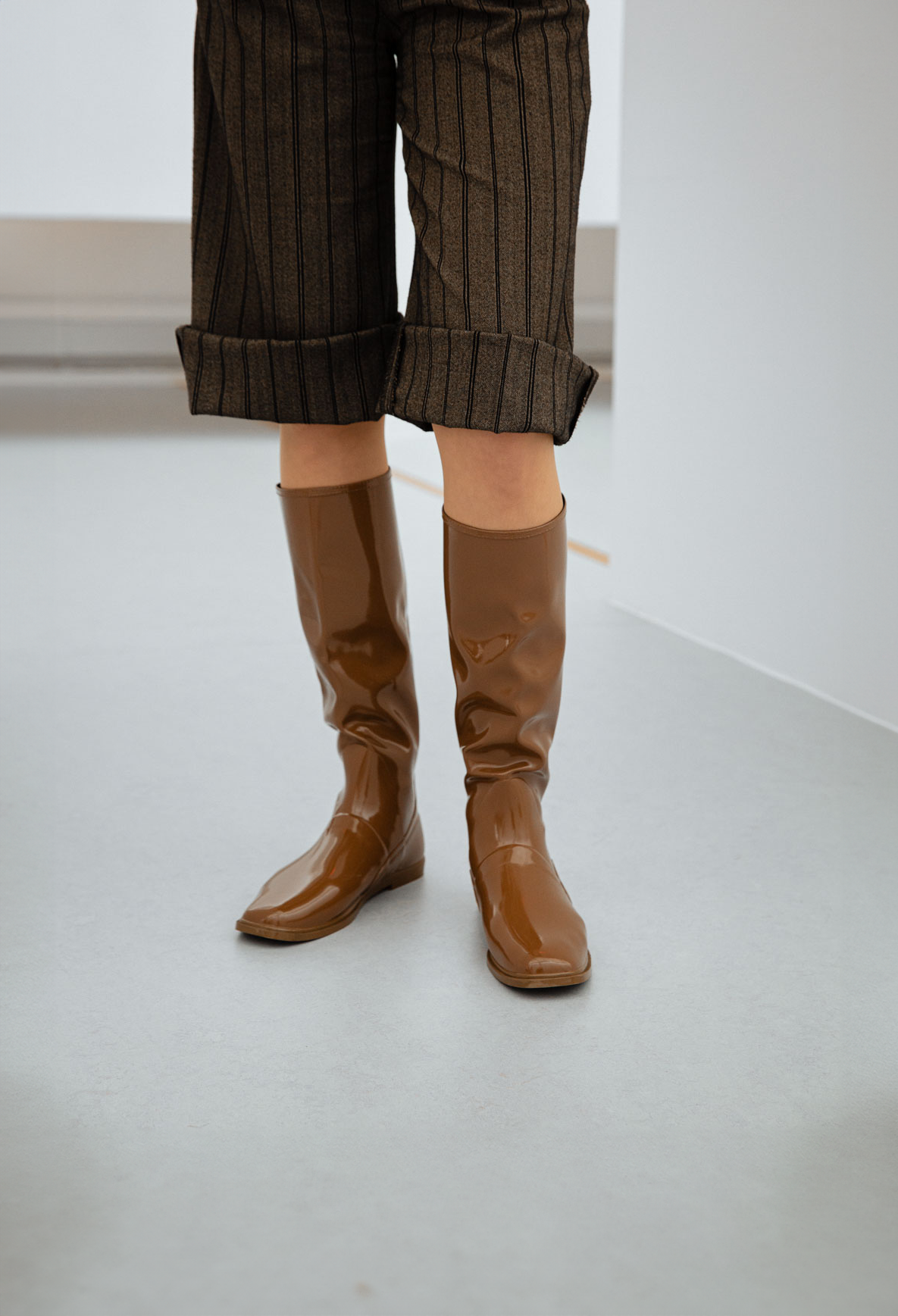 Close-up of Svea Walnut Brown knee-high boots by ACQUO of Sweden paired with tailored pinstripe trousers in a minimalist studio. Elegant waterproof boots with clean Swedish design, square toes and a modern understated silhouette in rich walnut brown.