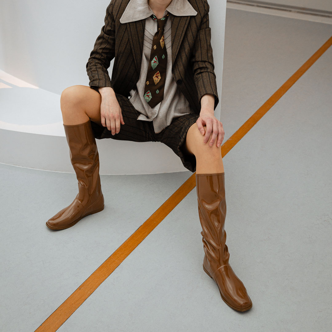 Woman wearing Svea Walnut Brown knee-high boots by ACQUO of Sweden styled with a tailored pinstripe suit and patterned tie in a contemporary studio setting. Waterproof boots with minimalist Swedish design, square toes and an elegant modern silhouette in warm walnut brown