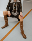 Woman wearing Svea Walnut Brown knee-high boots by ACQUO of Sweden styled with a tailored pinstripe suit and patterned tie in a contemporary studio setting. Waterproof boots with minimalist Swedish design, square toes and an elegant modern silhouette in warm walnut brown
