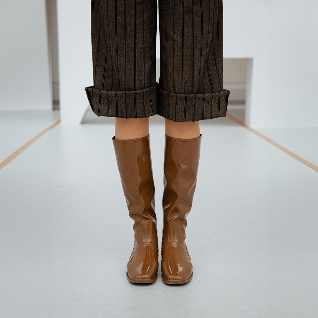 Front view of Svea Walnut Brown knee-high boots by ACQUO of Sweden paired with cropped pinstripe trousers in a minimalist studio interior. Elegant waterproof boots with clean Swedish design, square toes and a sleek contemporary silhouette in rich walnut brown.