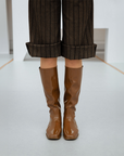 Front view of Svea Walnut Brown knee-high boots by ACQUO of Sweden paired with cropped pinstripe trousers in a minimalist studio interior. Elegant waterproof boots with clean Swedish design, square toes and a sleek contemporary silhouette in rich walnut brown.