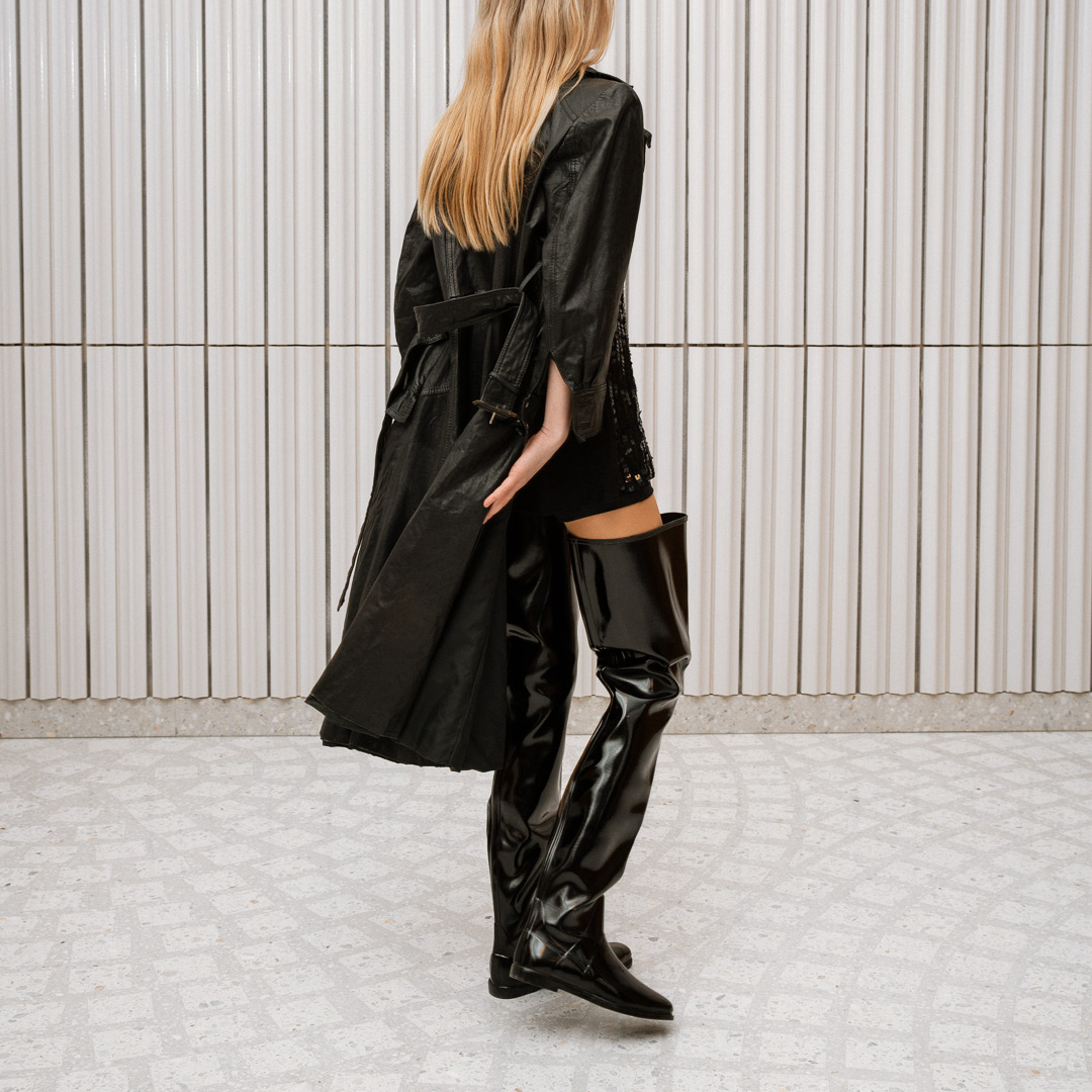 Model dressed in ACQUO of Sweden high-shine over-the-knee black boots, paired with a black embellished top, short bottoms, and a flowing black trench coat – a bold and elegant look set in a modern Scandinavian space