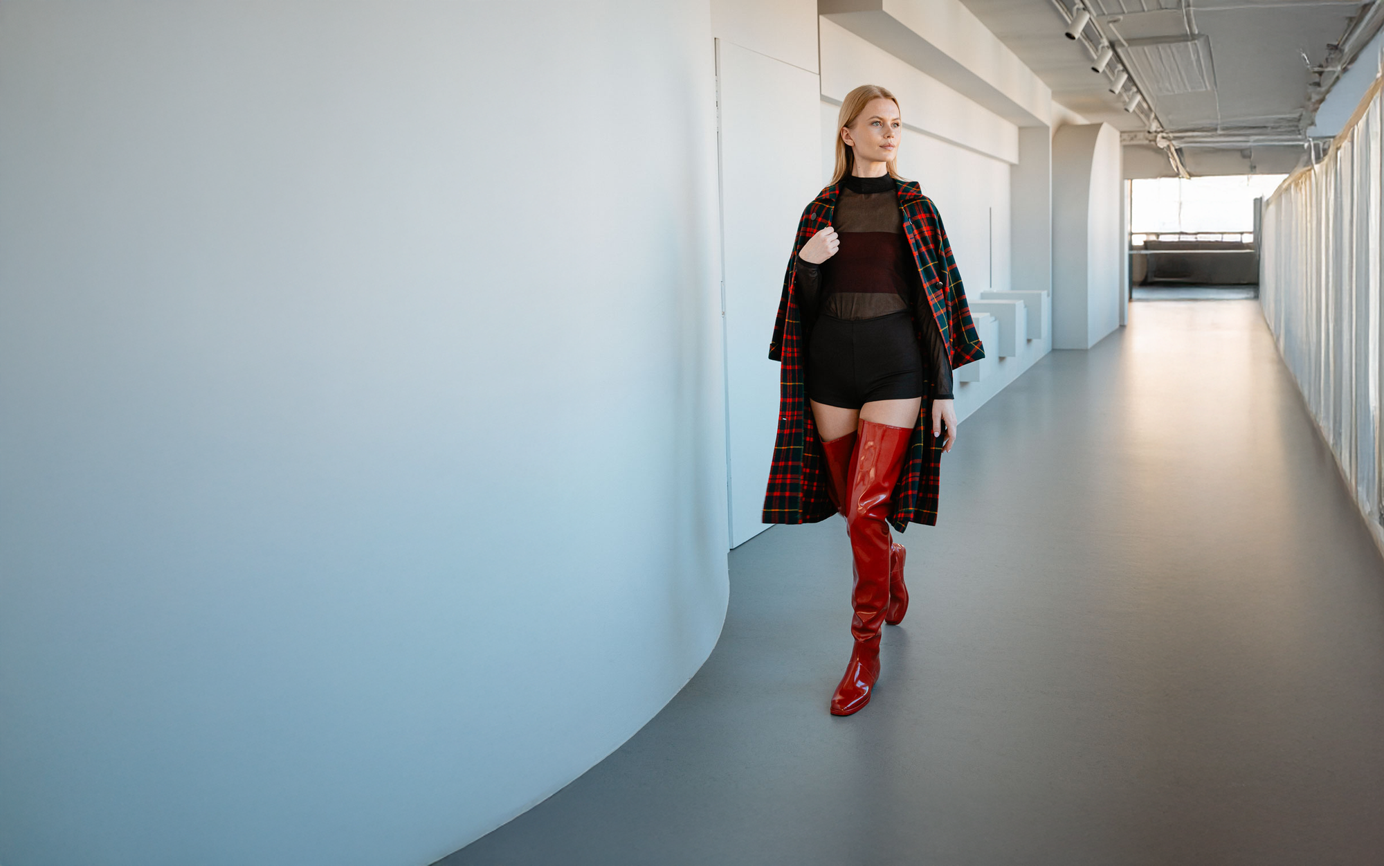 Woman walking through a minimalist interior wearing cherry red over-the-knee waterproof boots by ACQUO of Sweden. Contemporary Swedish design with a square toe, styled with a plaid coat and modern fashion silhouette