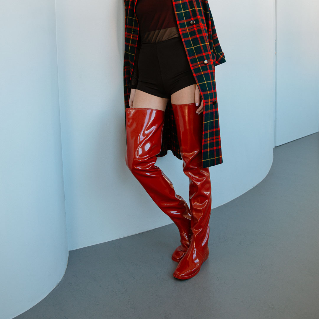 Model wearing Vega Cherry Red over-the-knee rubber boots by ACQUO of Sweden with a tartan coat and black shorts in a modern interior. Elegant waterproof statement boots featuring contemporary Swedish design and a sleek silhouette."