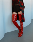 Model wearing Vega Cherry Red over-the-knee rubber boots by ACQUO of Sweden with a tartan coat and black shorts in a modern interior. Elegant waterproof statement boots featuring contemporary Swedish design and a sleek silhouette."