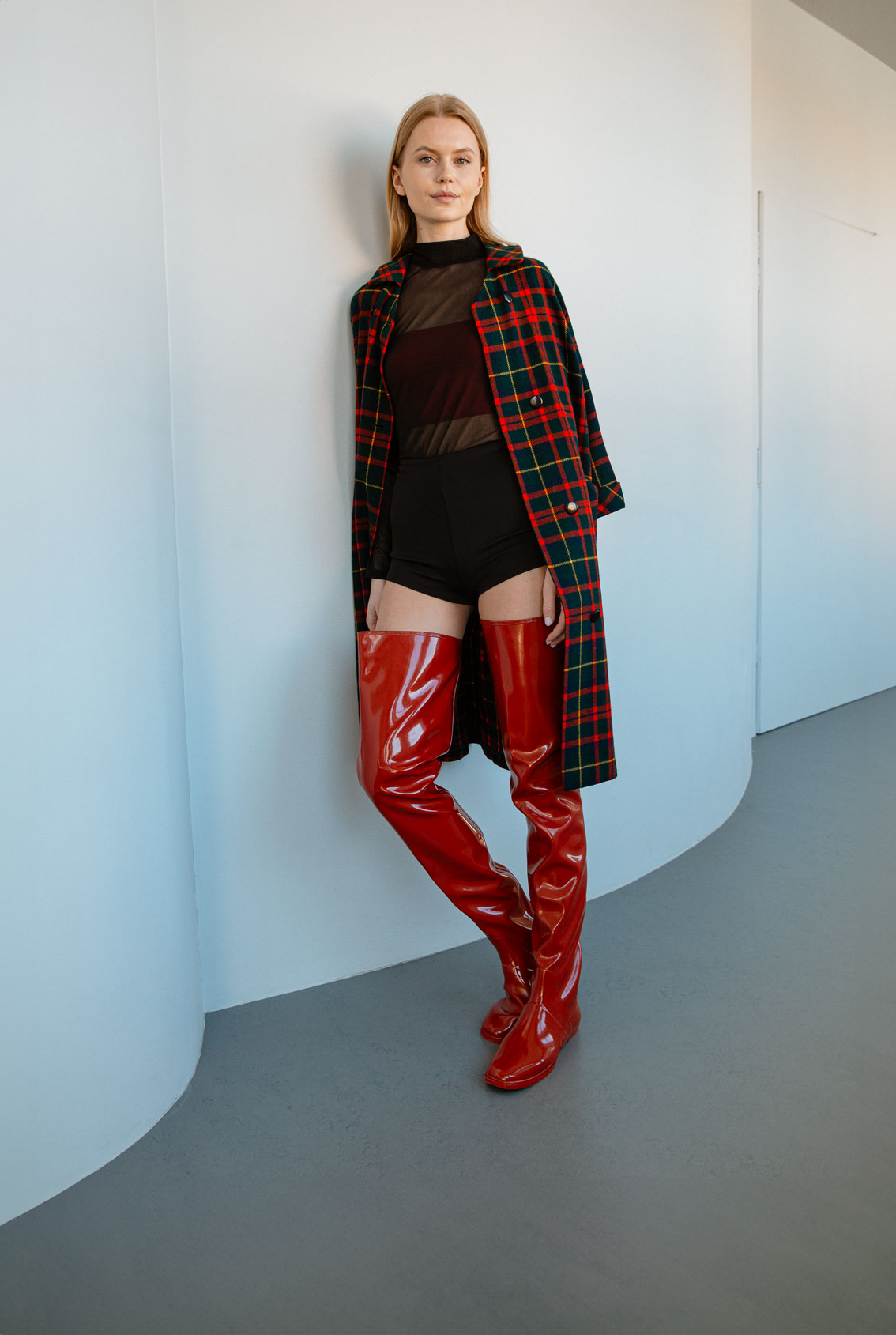 Model wearing Vega Cherry Red over-the-knee boots by ACQUO of Sweden styled with a tartan coat and black bodysuit in a minimalist interior. Elegant waterproof boots showcasing bold Swedish design and contemporary Scandinavian fashion