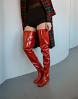 Model wearing Vega Cherry Red over-the-knee boots by ACQUO of Sweden styled with a tartan coat and black outfit in a minimalist indoor setting. Bold waterproof boots with elegant Swedish design and a modern Scandinavian silhouette.