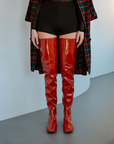 Model wearing Vega Cherry Red over-the-knee boots by ACQUO of Sweden with a tartan coat and black shorts in a minimalist interior. Statement waterproof boots featuring elegant Swedish design and a modern Scandinavian aesthetic