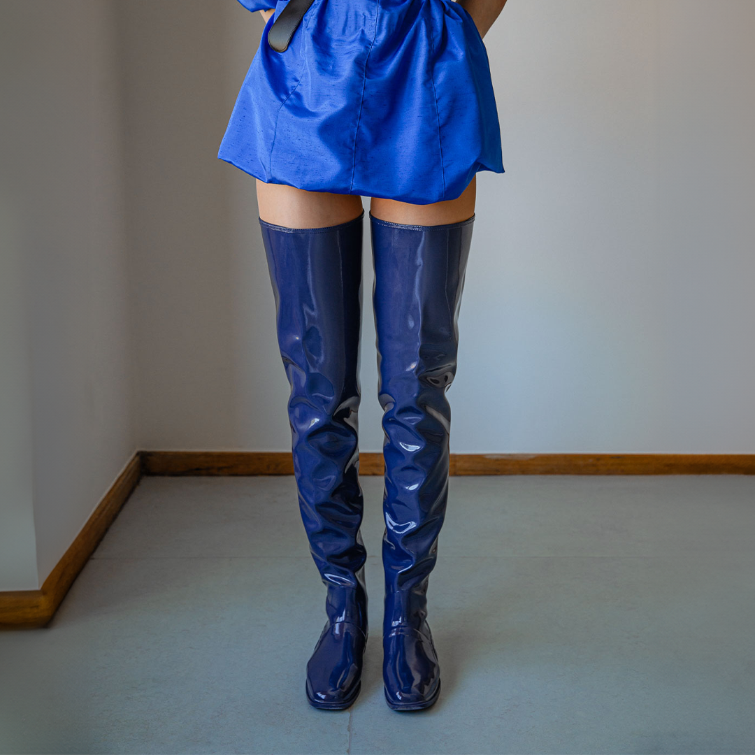 Front view of a model wearing Vega Iris Blue boots by ACQUO of Sweden. The image highlights elegant iris blue over-the-knee rubber boots with a sleek fitted silhouette, square toe, and reflective finish, styled with a vibrant blue mini dress in a softly lit modern interior with wooden floor details
