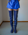 Front view of a model wearing Vega Iris Blue boots by ACQUO of Sweden. The image highlights elegant iris blue over-the-knee rubber boots with a sleek fitted silhouette, square toe, and reflective finish, styled with a vibrant blue mini dress in a softly lit modern interior with wooden floor details