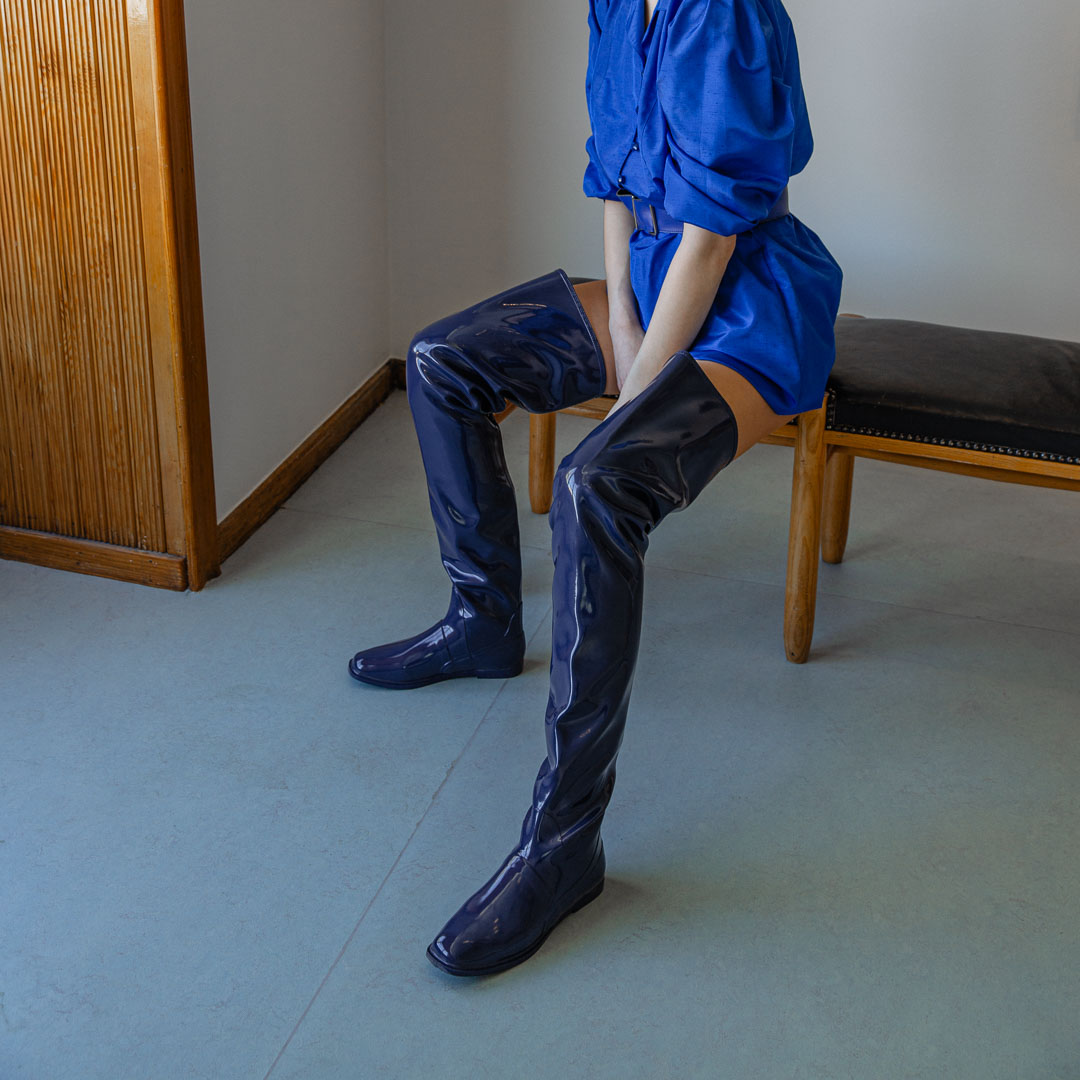 Model seated on a wooden bench wearing Vega Iris Blue boots by ACQUO of Sweden. The image highlights elegant iris blue over-the-knee rubber boots with a sleek fitted silhouette, square toe, and reflective finish, styled with a vibrant blue belted mini dress in a softly lit modern interior with wooden wall details.