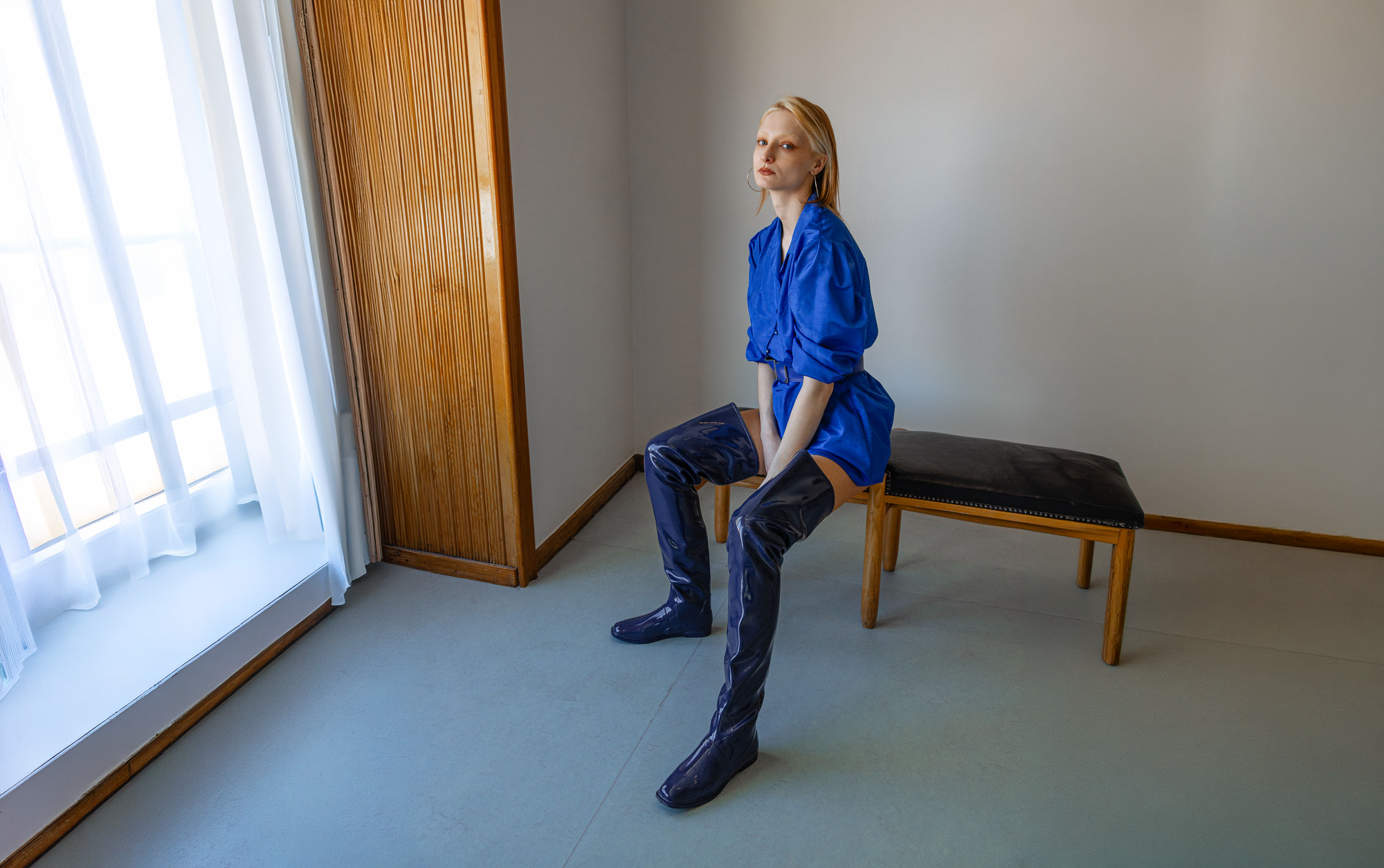 Model seated on a wooden bench wearing Vega Iris Blue boots by ACQUO of Sweden. The image highlights elegant iris blue over-the-knee rubber boots with a sleek fitted silhouette, square toe, and reflective finish, styled with a vibrant blue belted mini dress in a softly lit modern interior with wooden wall details.