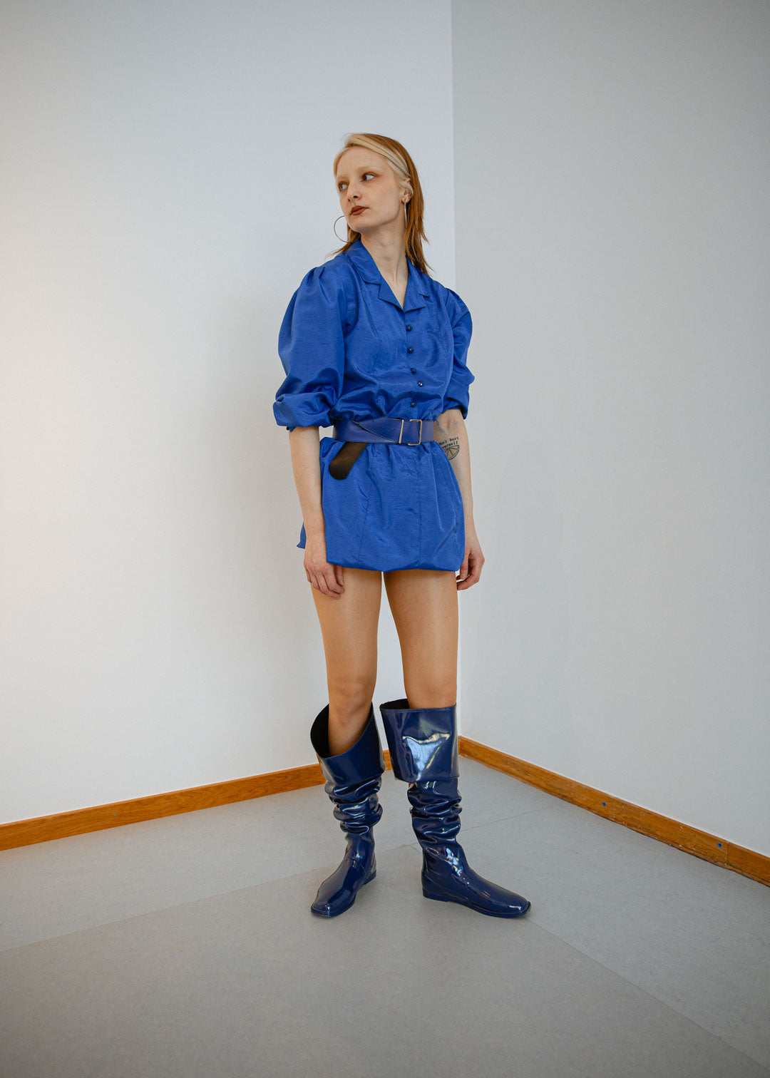 Full-length image of a model standing in a minimalist indoor space wearing Vega Iris Blue boots by ACQUO of Sweden. The iris blue rubber boots are styled folded down below the knees with a reflective finish and square toe silhouette, paired with a cobalt blue belted mini dress.