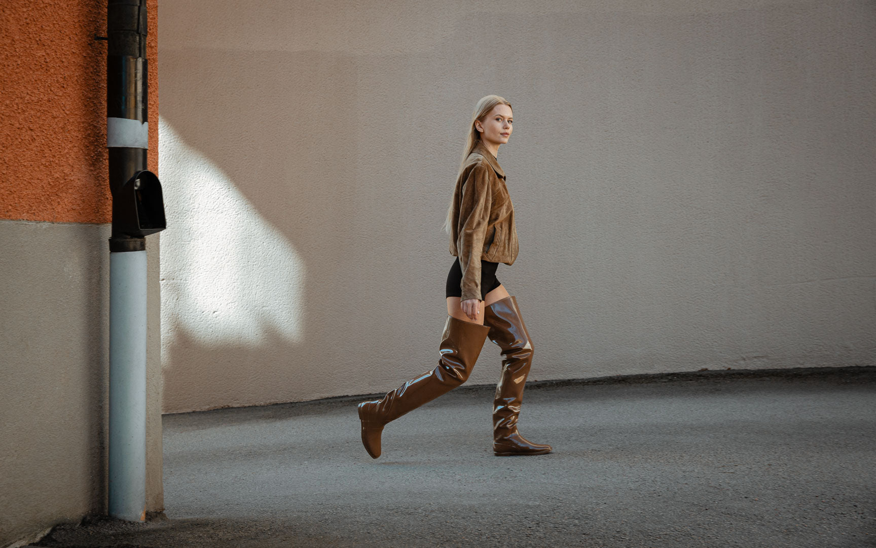 Woman walking outdoors in walnut brown over-the-knee waterproof boots by ACQUO of Sweden. Contemporary Swedish design with a square toe and sleek silhouette, styled with a brown jacket for a modern Scandinavian look